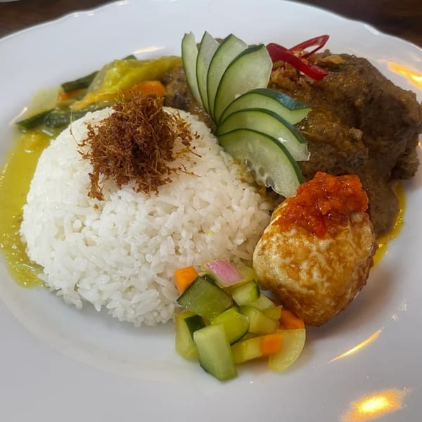 A white plate with rice, rendang, vegetables, cucumber slices, and a fried egg with chili sauce.